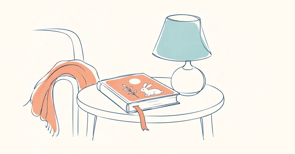 Closed book and lamp on a side table beside a chair draped with a blanket.